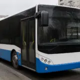 O fotografie a unui autobuz de transport public, vopsit în albastru și alb, parcat pe marginea unei străzi urbane lângă un bloc de apartamente