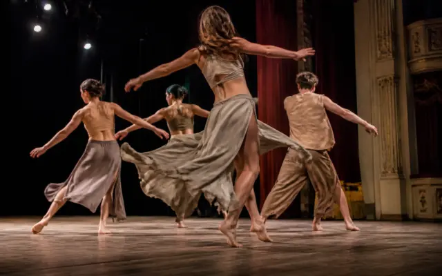 Patru dansatori sunt pe o scenă de teatru, surprinși în mișcare în timpul unui spectacol