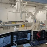O imagine dintr-o sală de control medicală care privește printr-un geam către o sală de operație sau intervenție dotată cu un angiograf modern cu braț C