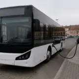 Un autobuz modern, alb cu negru, este parcat langa o statie de incarcare electrica alba intr-o zona urbana